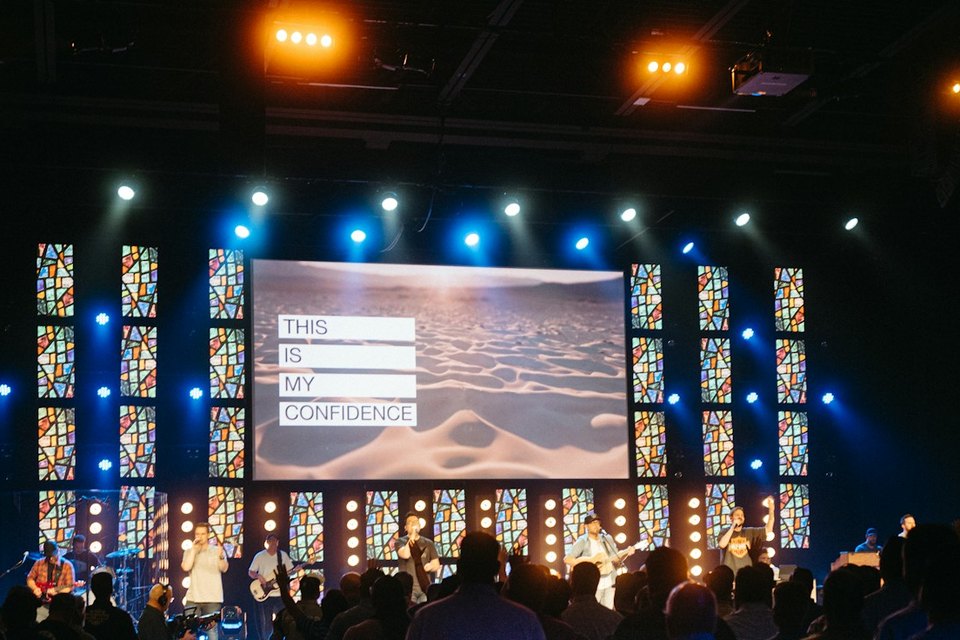 Song words on an LED wall between LED panels, the worship team lit on stage and the room standing to sing