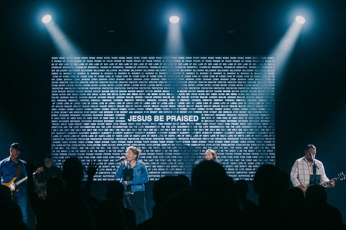 “Jesus Be Praised” on an LED wall behind the worship team, stage lights above